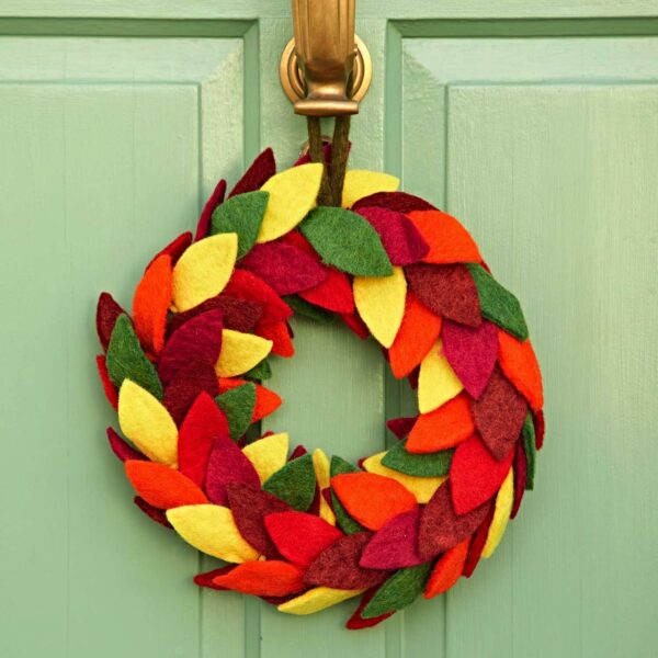 The Felt Colourful Leaf Wreath, featuring layered red, yellow, green, and orange felt leaves, hangs on a light green door with a brass knocker.