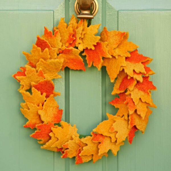 The Felt Autumn Leaves Wreath, featuring vibrant orange and yellow felt leaves, hangs beautifully on a green door.