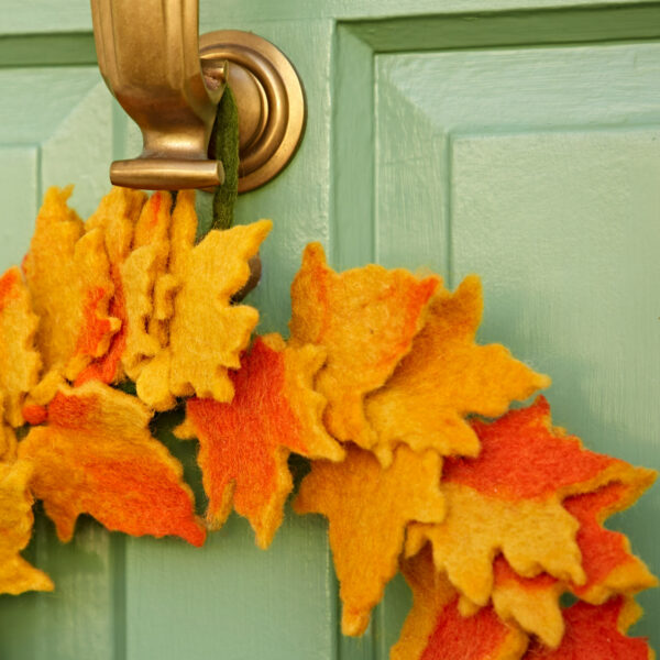 A close-up of a brass door knocker and the Felt Autumn Leaves Wreath, featuring yellow and orange tones, displayed on a light green door.