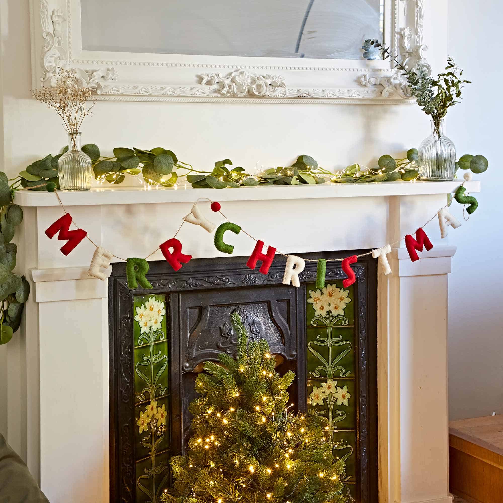 A Felt Merry Christmas Letter Garland decorates a white mantel with greenery, hanging above a lit Christmas tree and tiled fireplace.