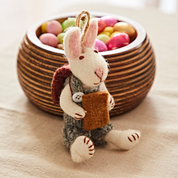 A Felt Rabbit with Journal and Rucksack Hanging Decoration sits in front of a wooden bowl of colourful candy-coated chocolates and holds a brown felt piece.