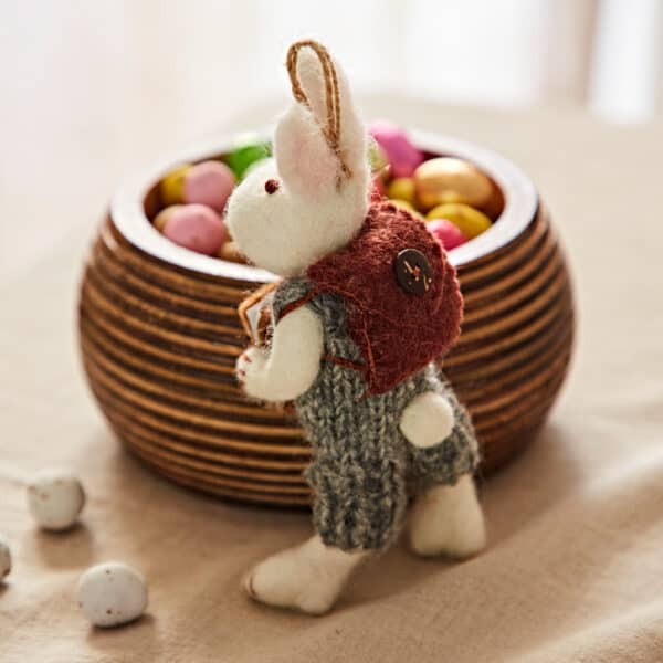 The Felt Rabbit with Journal and Rucksack Hanging Decoration, dressed in a red waistcoat and grey trousers, stands beside a wooden bowl of colourful sweet eggs on a beige surface.