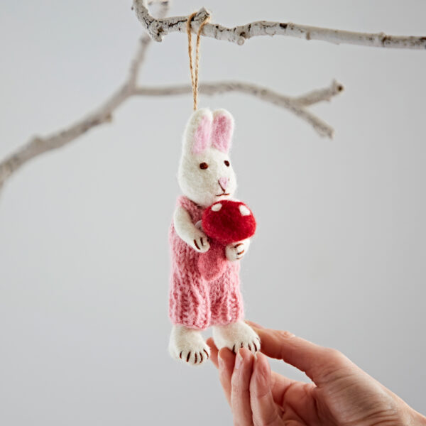 A hand holds the Felt Rabbit with Mushroom Hanging Decoration—a rabbit in pink dungarees holding a red and white toadstool—hanging from a bare tree branch.
