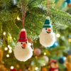 Two Felt Snowman Pom Pom Baubles, each wearing red and blue hats, hang on a Christmas tree adorned with festive lights, baubles, and decorations.