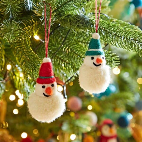 Two Felt Snowman Pom Pom Baubles, each wearing red and blue hats, hang on a Christmas tree adorned with festive lights, baubles, and decorations.