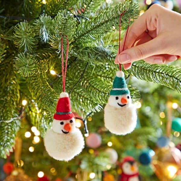 A hand hangs the Felt Snowman Pom Pom Bauble with a blue hat next to another snowman ornament with a red hat on a decorated Christmas tree.