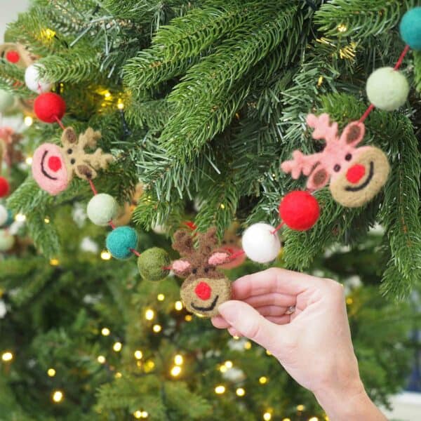 A hand hangs a Felt Reindeer Garland on a decorated Christmas tree with lush green branches and sparkling fairy lights.