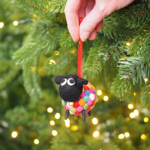 A hand hangs the Felt Sheep Ornament Christmas Decoration with a red ribbon on a Christmas tree, adding charm to the festive décor and twinkling fairy lights.