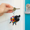 A hand holds a key with the Felt Sheep Keyring, made from colourful felt balls, near a blue door with a silver lock.