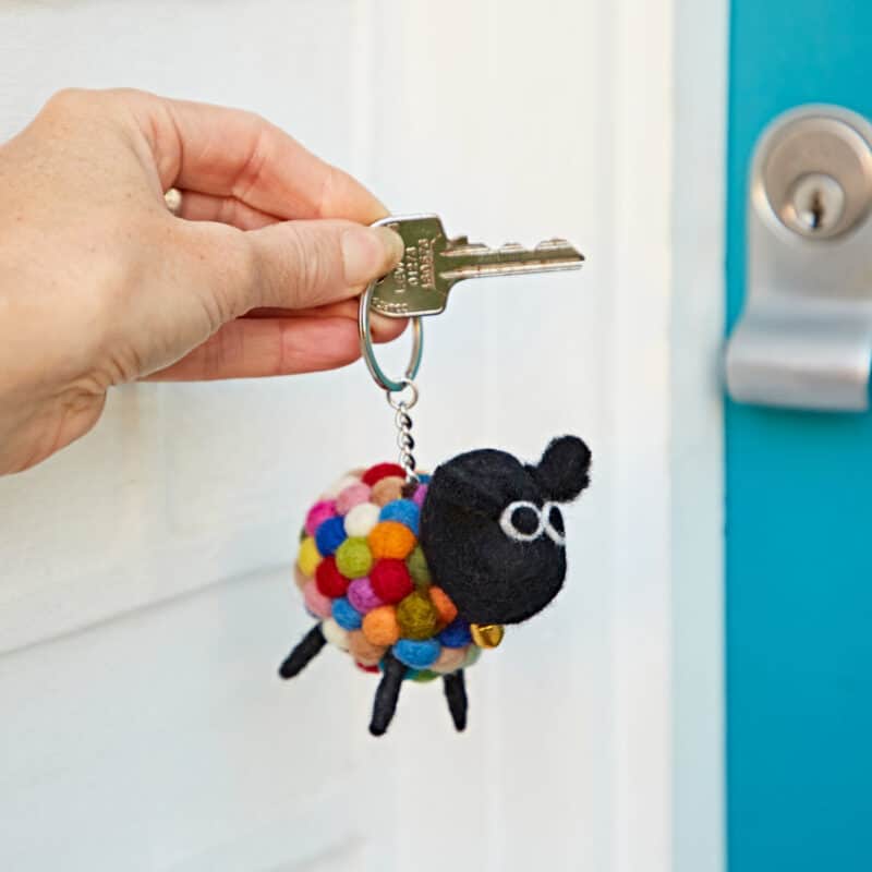 A hand holds a key with the Felt Sheep Keyring, made from colourful felt balls, near a blue door with a silver lock.