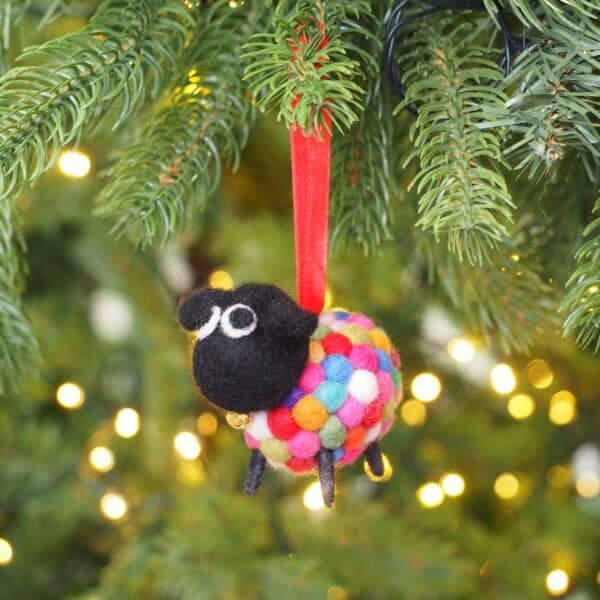 The Felt Sheep Ornament Christmas Decoration, featuring a colourful body and black head, hangs from a red ribbon as a festive accent on a Christmas tree with twinkling lights in the background.