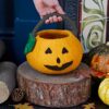 A hand holds a Felt Trick or Treat Halloween Pumpkin Basket with a jack-o'-lantern face, set on a tree stump surrounded by autumn leaves, logs, and pumpkins.