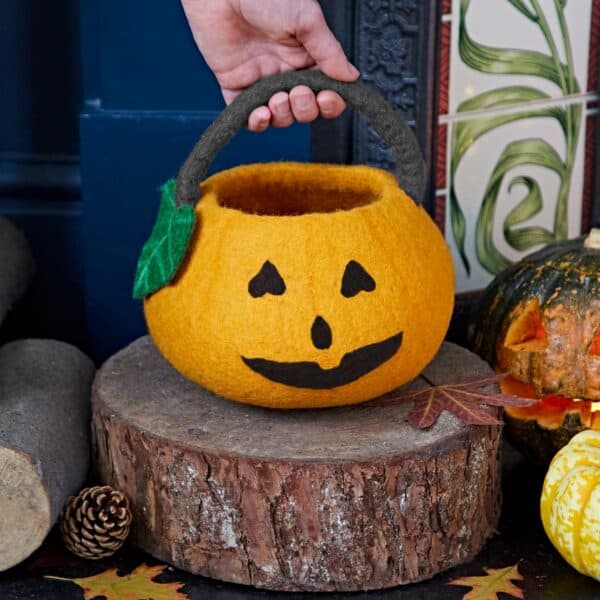 A hand holds a Felt Trick or Treat Halloween Pumpkin Basket with a jack-o'-lantern face, set on a tree stump surrounded by autumn leaves, logs, and pumpkins.