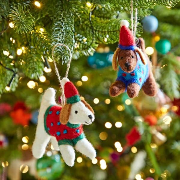 Two Felt Dog in Jumper Christmas Decorations, each wearing a red hat and festive jumper, hang on the Christmas tree amid twinkling lights and lush green branches.
