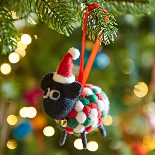 The Felt Sheep in Santa Hat Christmas Decoration, featuring a red ribbon, hangs on a tree branch amid softly blurred colourful lights in the background.