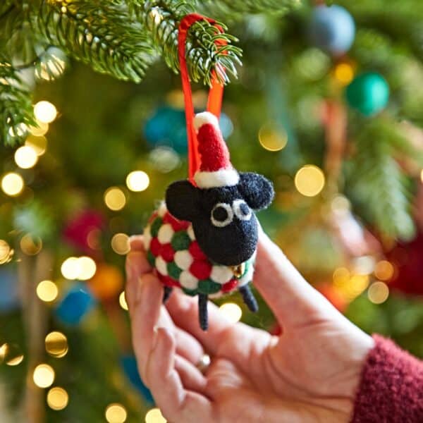 A hand holds the Felt Sheep in Father Christmas Hat Christmas Decoration, with a softly blurred, decorated and lit Christmas tree in the background.