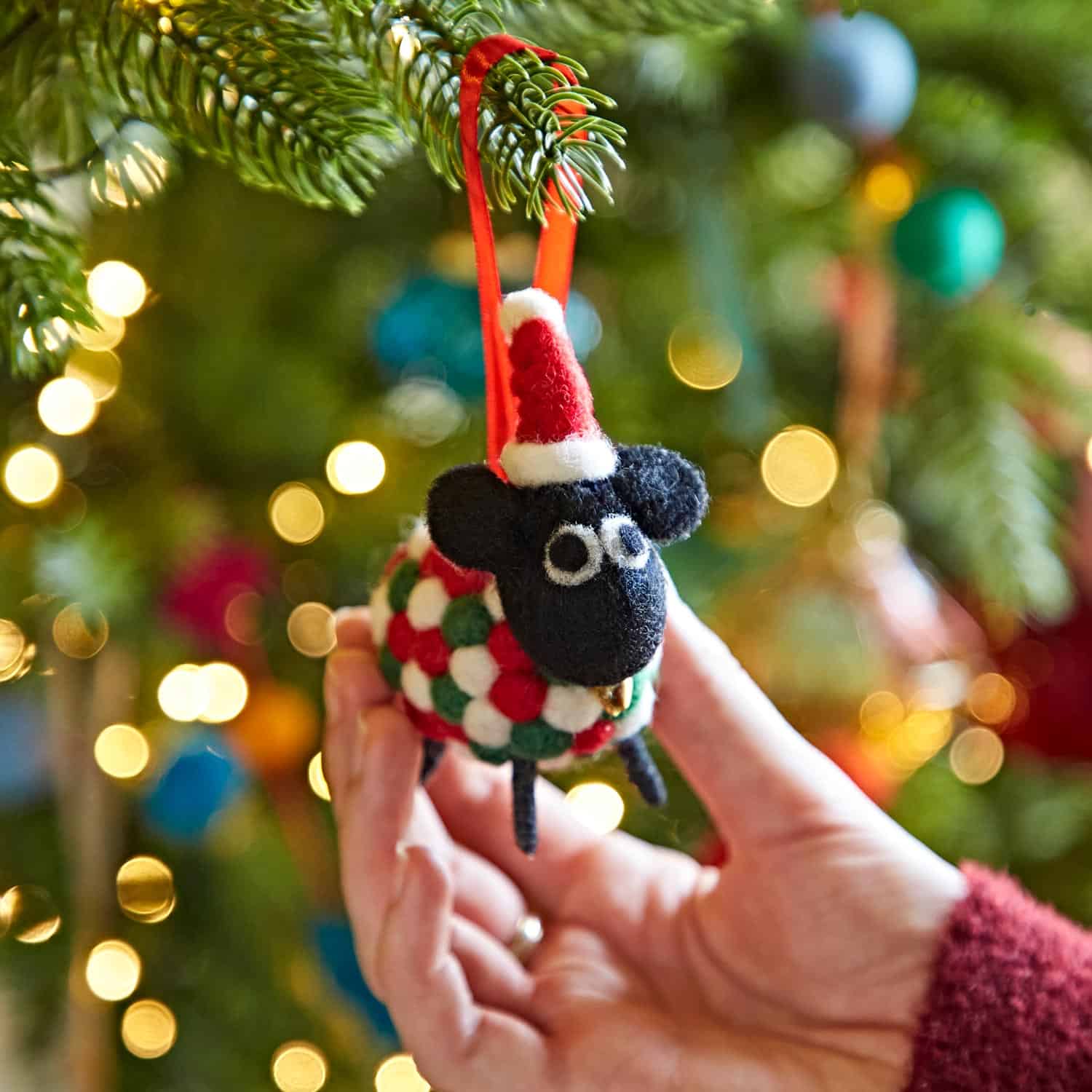 A hand holds the Felt Sheep in Father Christmas Hat Christmas Decoration, with a softly blurred, decorated and lit Christmas tree in the background.