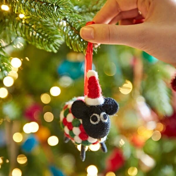 A hand hangs the Felt Sheep in Father Christmas Hat Christmas Decoration on a festive Christmas tree sparkling with twinkling lights and colourful baubles.