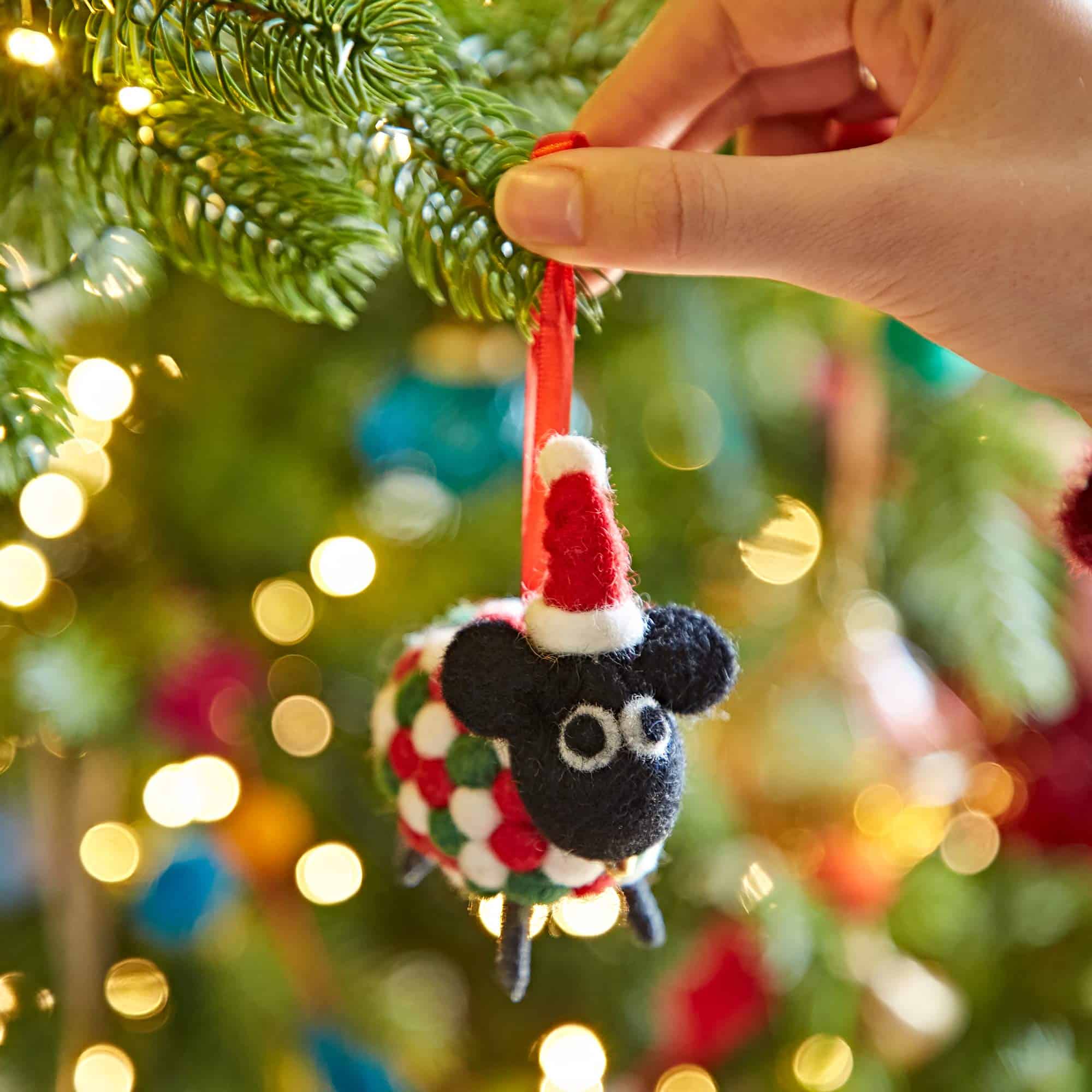 A hand hangs the Felt Sheep in Father Christmas Hat Christmas Decoration on a festive Christmas tree sparkling with twinkling lights and colourful baubles.