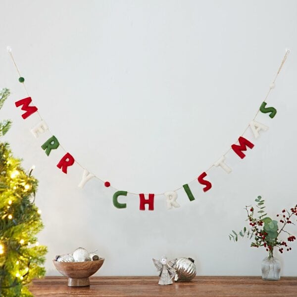 The Felt Merry Christmas Letter Garland is displayed on a white wall above a wooden table decorated with ornaments, greenery, and festive Christmas accents.