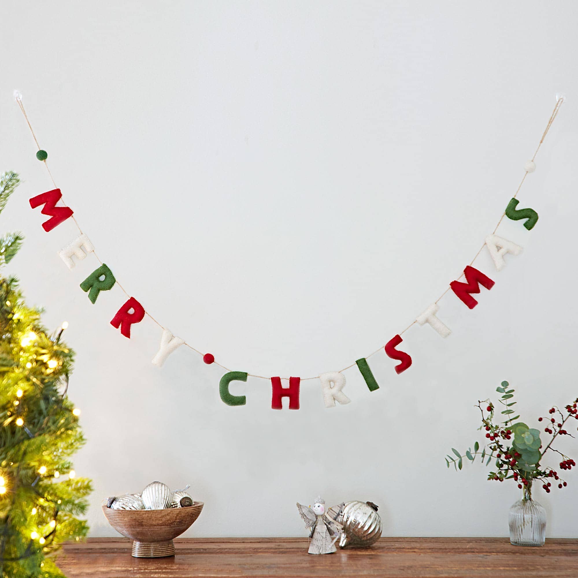 The Felt Merry Christmas Letter Garland is displayed on a white wall above a wooden table decorated with ornaments, greenery, and festive Christmas accents.