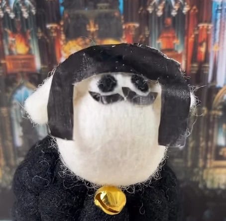 A handmade wool figure with black paper hair, black oval eyes, and a gold bell, set against a blurred cathedral-like backdrop—an ideal choice for Secret Santa Gifts Under £15.