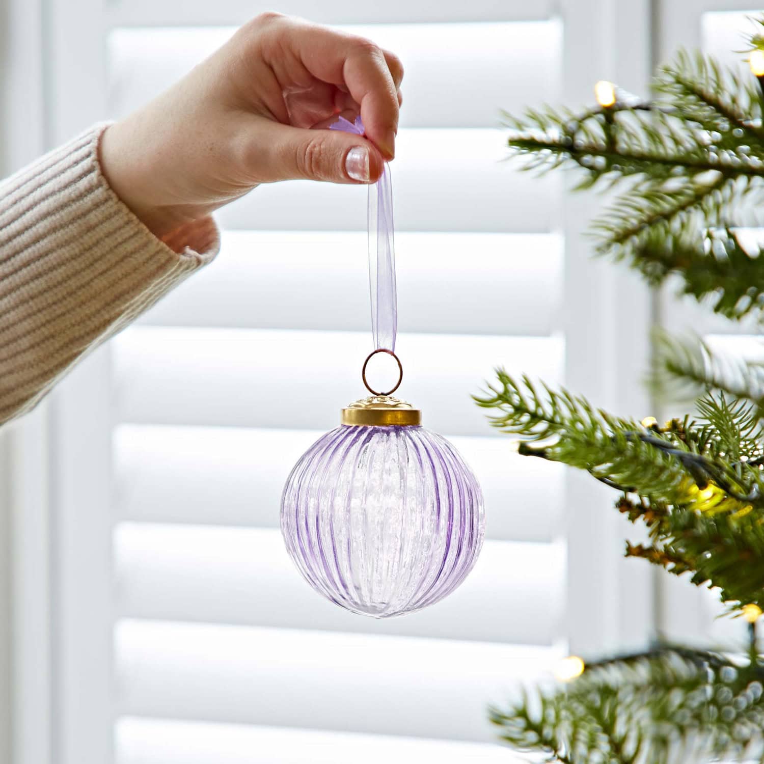 A hand holds a purple Christmas bauble crafted from recycled glass by a ribbon, preparing to hang it on a decorated tree.