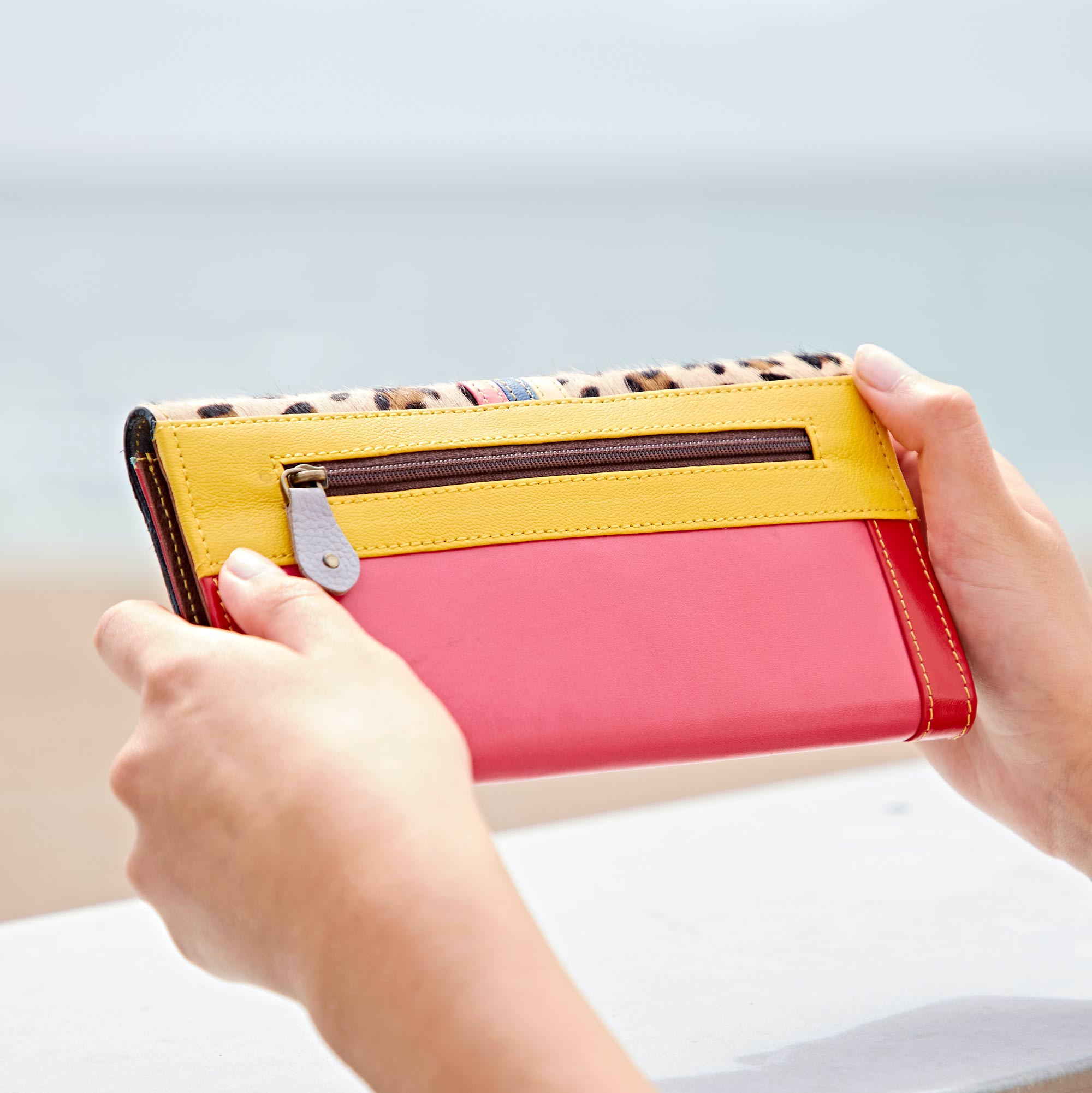 A person holds the Hema Multicoloured Recycled Leather Clutch Purse, featuring a vibrant yellow and pink design with a zip-fastened pocket, against a blurred outdoor background.