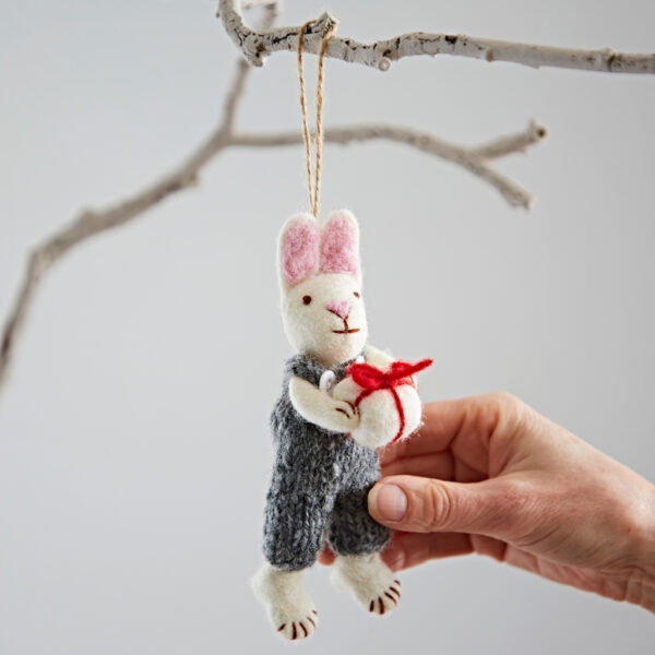 A hand displays the Felt Rabbit with Gift Hanging Decoration, featuring a grey outfit and red present, hanging by a string from a white branch.