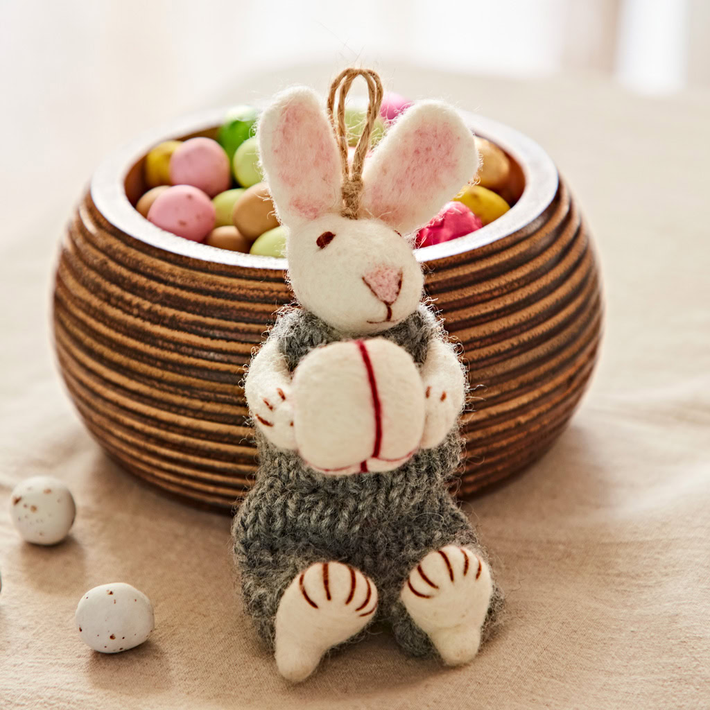 The Felt Rabbit with Gift Hanging Decoration holding an egg is displayed in front of a wooden bowl containing colourful candy-coated chocolates, all set on a beige surface.