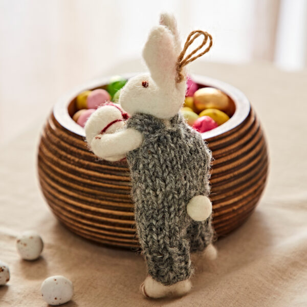 A Felt Rabbit with Gift Hanging Decoration in a grey knitted outfit stands beside a wooden bowl of colourful sweet eggs on a beige cloth.