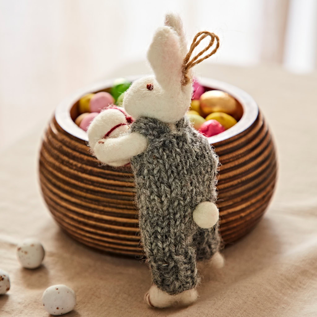 A Felt Rabbit with Gift Hanging Decoration in a grey knitted outfit stands beside a wooden bowl of colourful sweet eggs on a beige cloth.