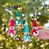 A hand adjusts one bauble from the Pack of 4 Colourful Felt Snowmen Christmas Decorations hanging on a Christmas tree, each snowman featuring bright hats and scarves with soft, glowing lights in the background.