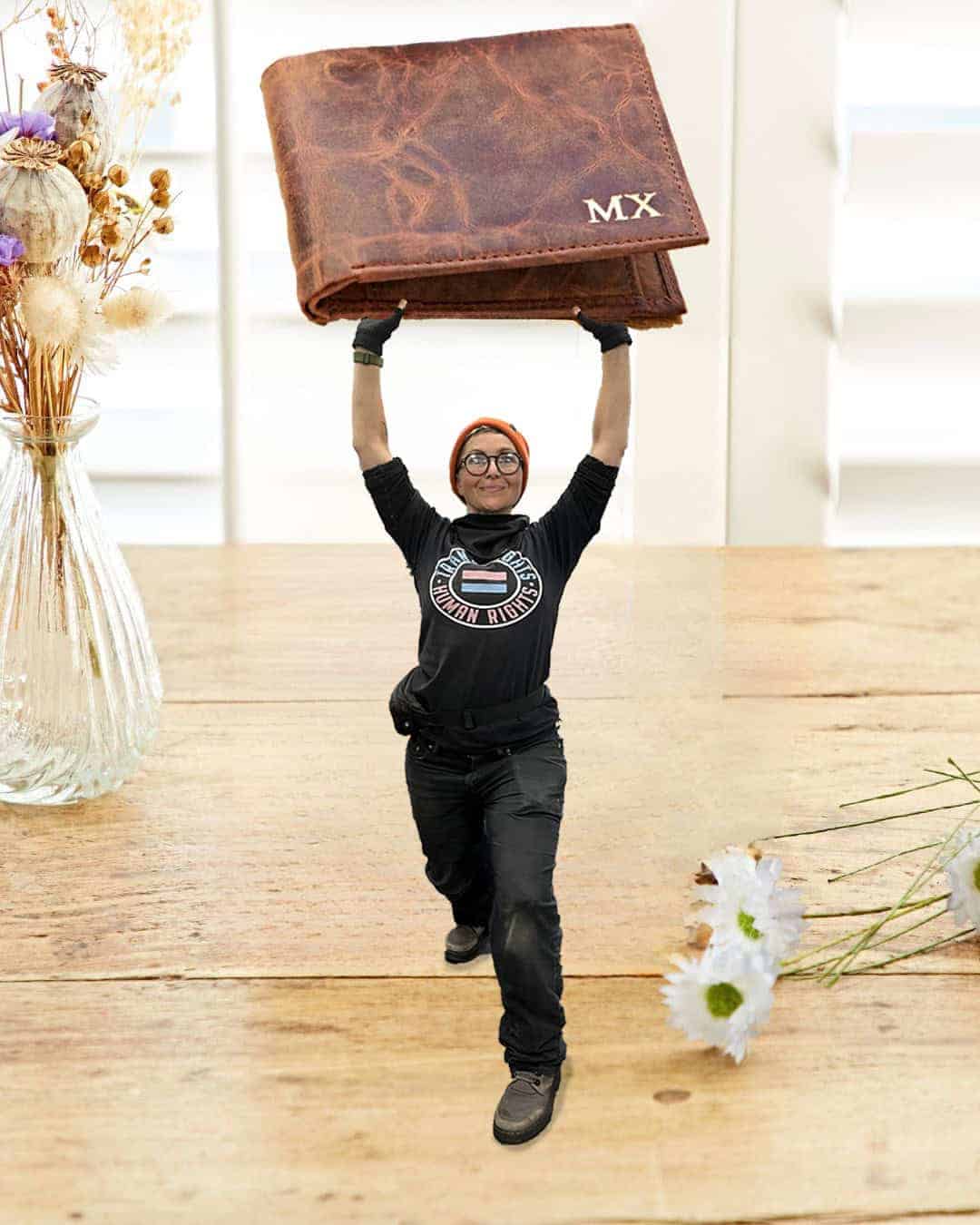 A person holding an oversized brown wallet marked with "MX" stands on a wooden table, surrounded by vases of dried and fresh flowers—one of our Staff Favourites for Christmas.