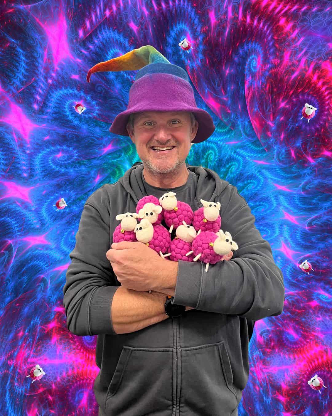 A smiling man in a purple hat holds several plush pink sheep against a vibrant, psychedelic blue and pink background—one of our Staff Favourites for Christmas.