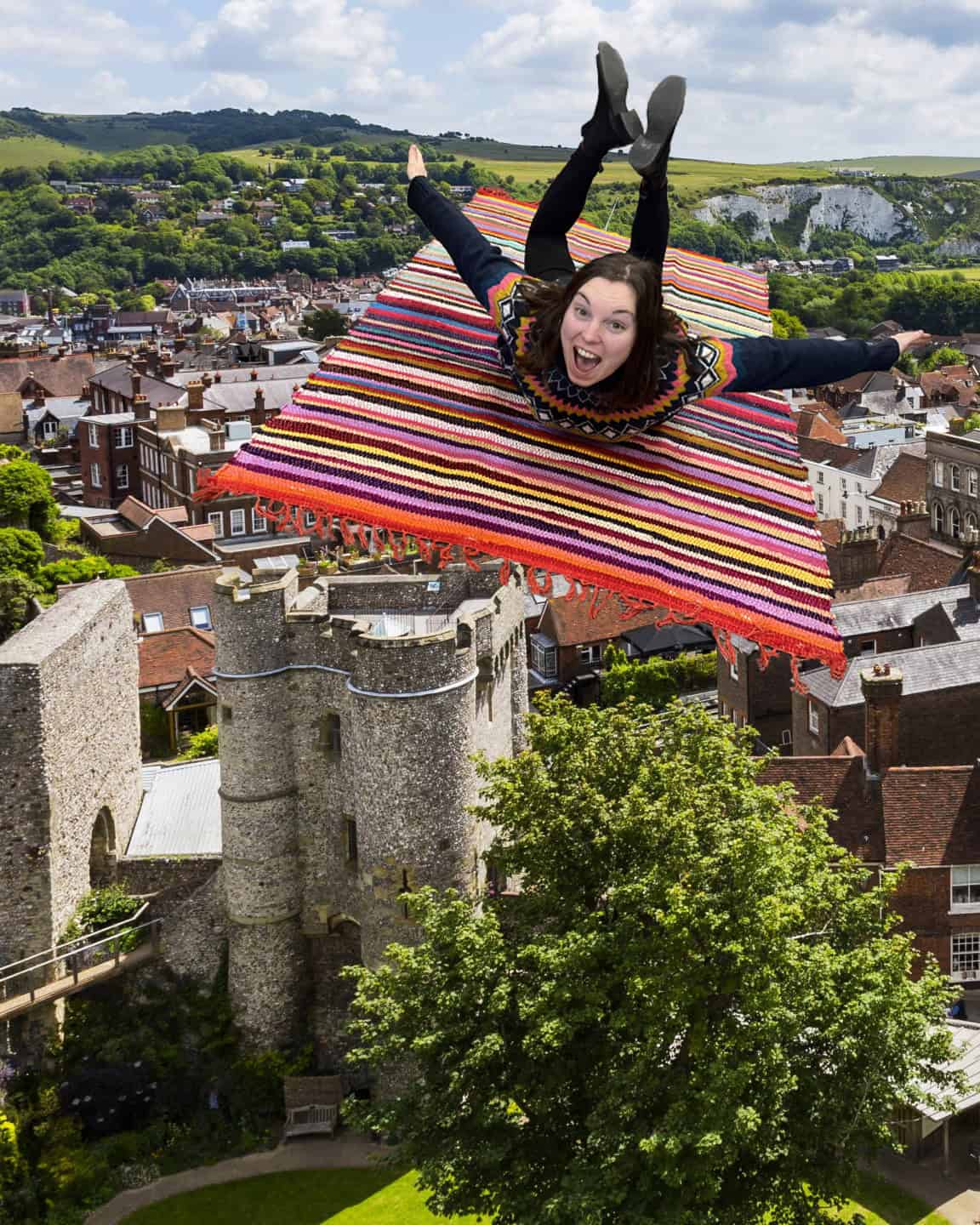A woman smiles and poses as she appears to fly on a striped magic carpet above a historic town with stone buildings and greenery—a scene among our Staff Favourites for Christmas.