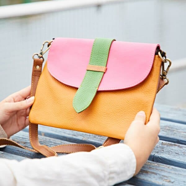 Person holding a multicoloured crossbody bag crafted from recycled leather, featuring a pink flap, green strap, and yellow body, placed on a wooden surface.