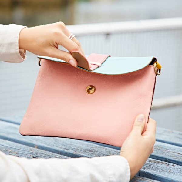 Someone opens the Priya Recycled Leather Multicoloured Crossbody Bag, featuring a gold clasp, whilst sitting outdoors on a blue wooden bench.