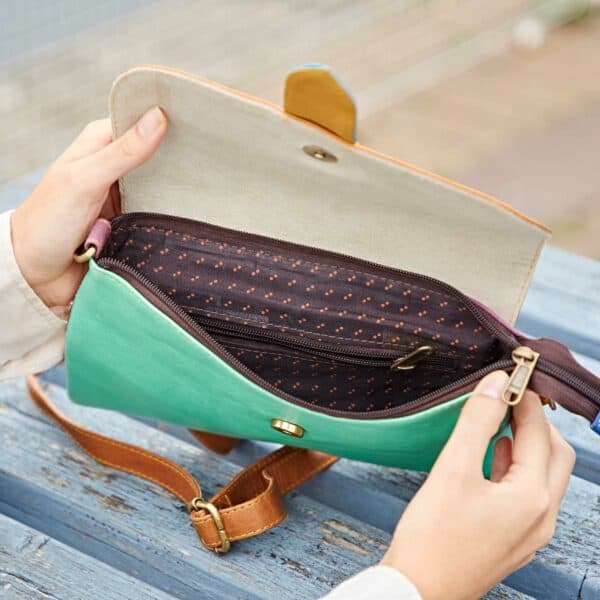Someone opens a Priya Recycled Leather Multicoloured Crossbody Bag, revealing a patterned inner zip pocket while sitting on a blue wooden surface.