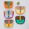 Six Seema Multicoloured Recycled Leather Crossbody Bags with vibrant bodies and patterned flaps are displayed in two columns on a light surface, topped with a small succulent.