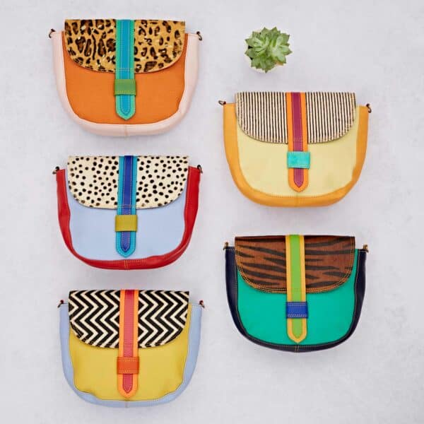 Six Seema Multicoloured Recycled Leather Crossbody Bags with vibrant bodies and patterned flaps are displayed in two columns on a light surface, topped with a small succulent.