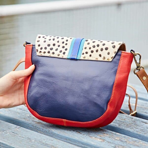 A hand holds the Seema Multicoloured Recycled Leather Crossbody Bag, featuring a navy body, red sides, brown strap, and a flap with black spots on white plus a blue stripe.