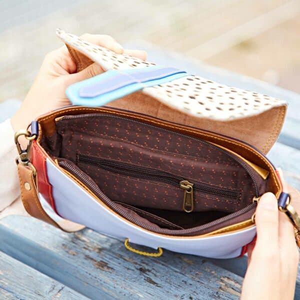 A person holds open the Seema Multicoloured Recycled Leather Crossbody Bag, showing a patterned flap, brown zip-fastened inner pocket, and matching patterned lining.