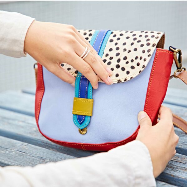 A person opens the Seema Multicoloured Recycled Leather Crossbody Bag, which features red sides, a blue strap, and a bold black-and-white spotted flap.