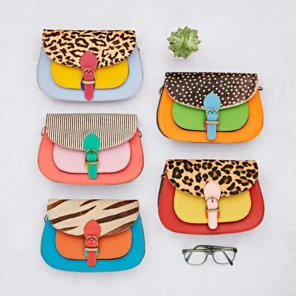 Six Sonashki Multicoloured Rectangular Saddle Bags with animal print flaps are arranged in two rows on a light surface, accompanied by a small succulent plant and spectacles.