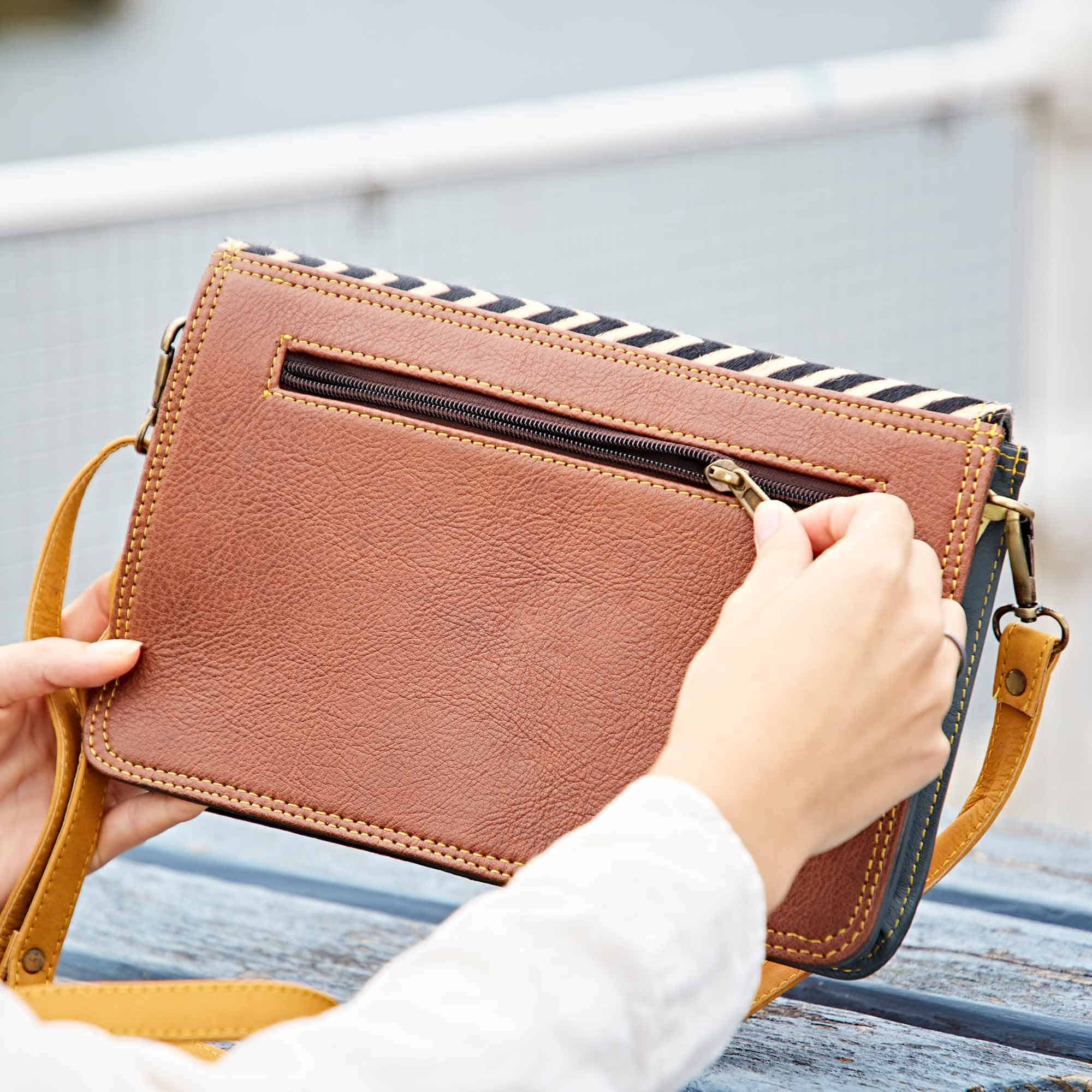 A person holds a Multicoloured Recycled Leather Crossbody Buckle Bag and unzips the back pocket whilst sitting outdoors near a metal fence.