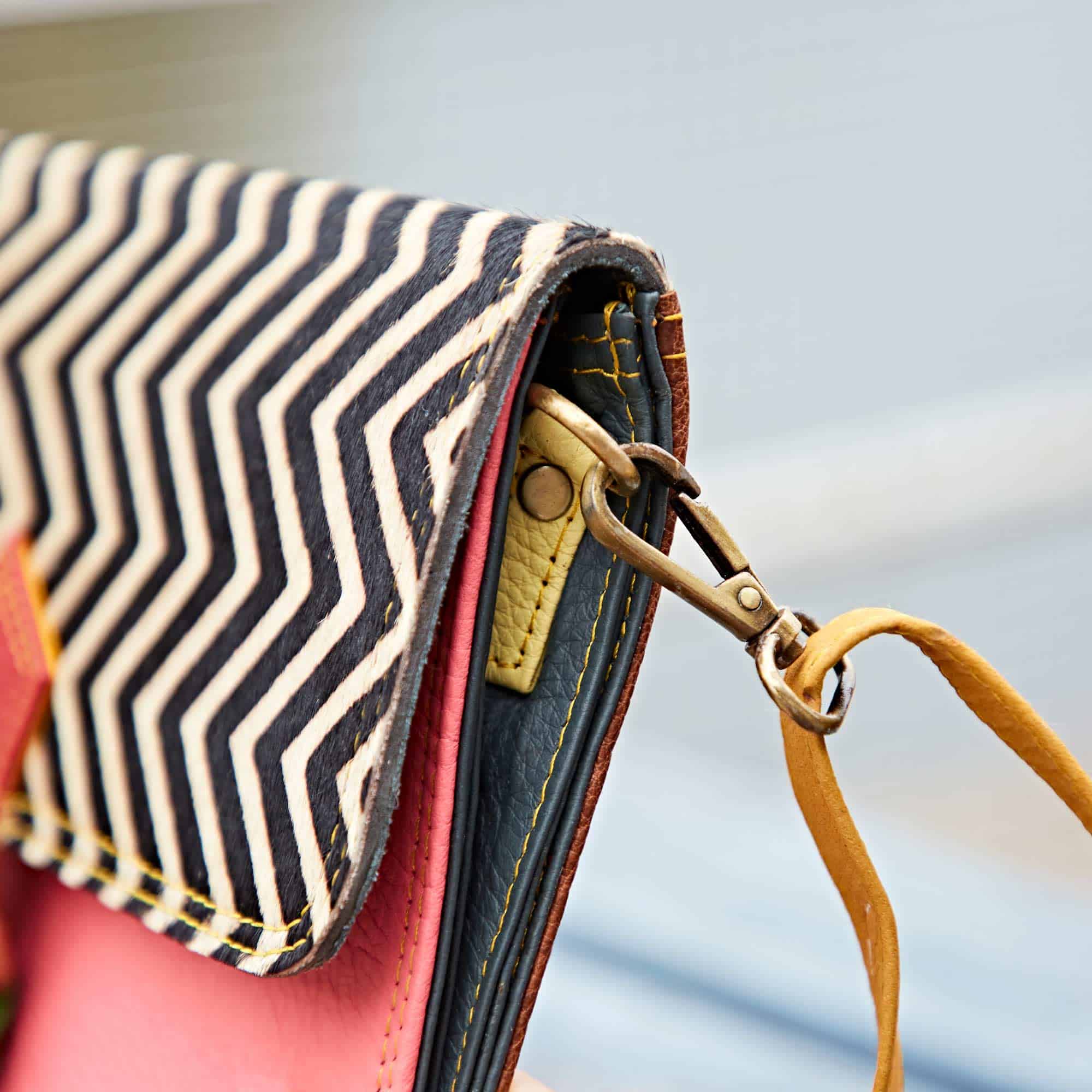 Close-up of the Multicoloured Recycled Leather Crossbody Buckle Bag, showcasing a black and white zigzag flap, pink body, yellow stitching, metal clasp, and a tan wrist strap.