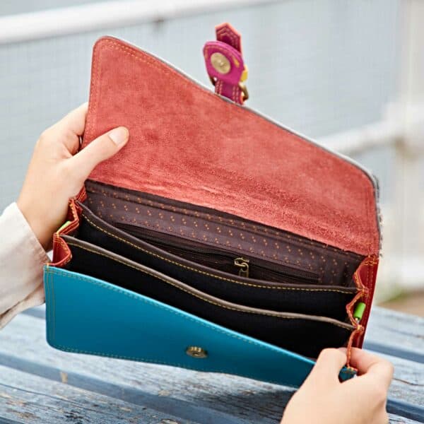 A person holds open the Multicoloured Recycled Leather Crossbody Buckle Bag, showing its multiple zipped and open compartments, on an outdoor surface.