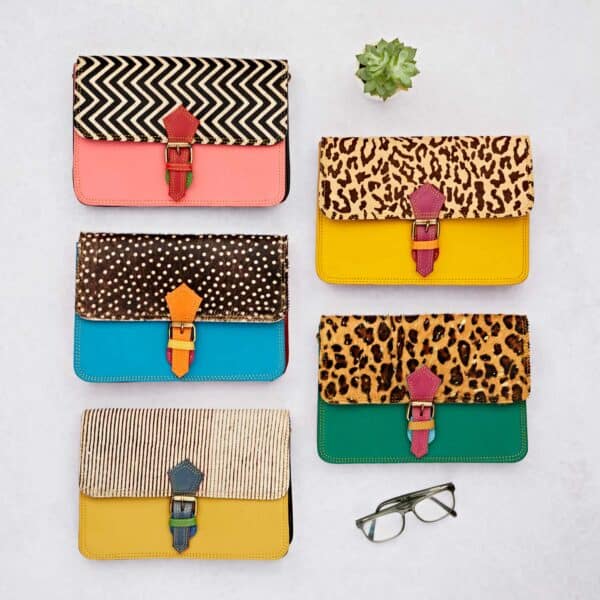 Six Multicoloured Recycled Leather Crossbody Buckle Bags with animal print and geometric patterns are displayed on a white surface next to a succulent plant and spectacles.