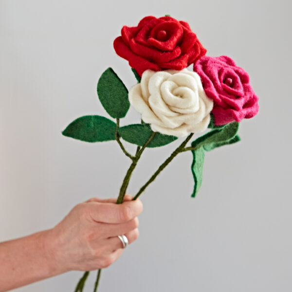 A hand holds three Felt Roses—red, pink, and white—with green felt leaves and stems against a plain light background.